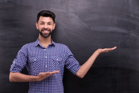 Photo of handsome young casual man on blank chalkboard background. Man smiling and cheerfully pointing at something with two handsの写真素材