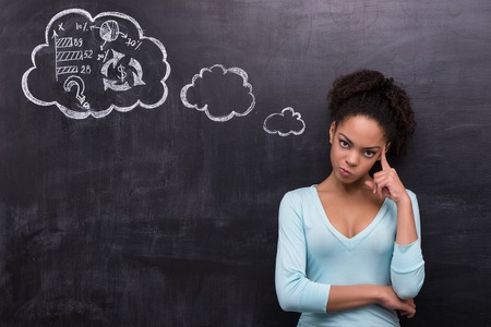 Photo of young dissatisfied afro-american woman on chalkboard background. Woman looking at camera and trying to solve a problem painted on chalkboardの写真素材