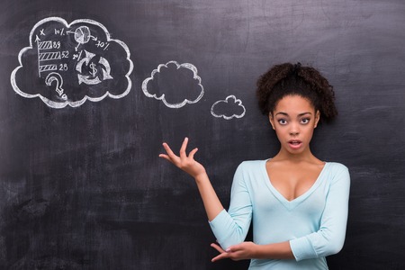 Photo of young surprised afro-american woman on chalkboard background. Woman looking at camera and trying to solve a problem painted on chalkboardの写真素材
