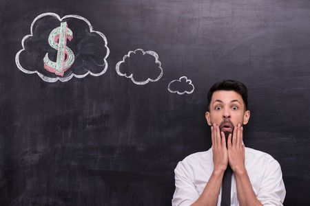 Photo of handsome young businessman on chalkboard background. Man anxiously looking at camera. Cloud formed dialog and dollar sign in it painted on chalkboardの写真素材