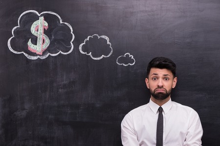 Photo of handsome young businessman on chalkboard background. Man sadly looking at camera. Cloud formed dialog and dollar sign in it painted on chalkboardの写真素材