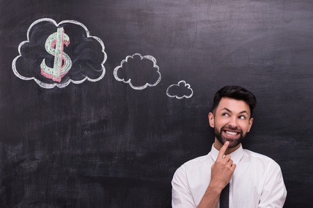 Photo of handsome young businessman on chalkboard background. Man cheerfully looking at cloud formed dialog and dollar sign in it painted on chalkboardの写真素材