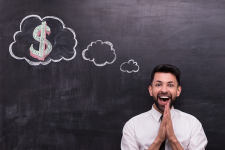 Photo of handsome young businessman on chalkboard background. Man very happily looking at camera. Cloud formed dialog and dollar sign in it painted on chalkboardの写真素材