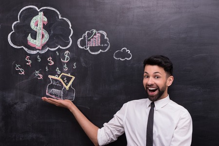 Photo of handsome young businessman on chalkboard background. Man smiling and cheerfully looking at camera. Dollar signs and purse painted on chalkboardの写真素材