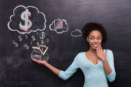 Photo of young afro-american woman on chalkboard background. Woman smiling and cheerfully looking at dollar signs and purse painted on chalkboardの写真素材