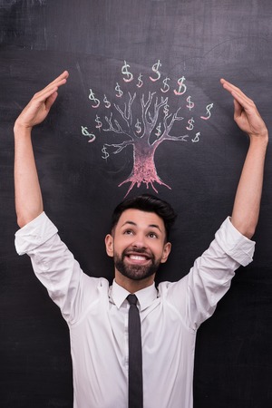 Photo of young businessman on chalkboard background. Man smiling and standing near dollar tree painted on chalkboardの写真素材