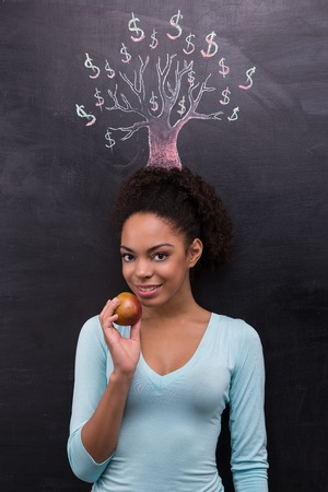 Photo of young afro-american woman on chalkboard background. Woman looking at camera and holding apple near dollar tree painted on chalkboardの写真素材