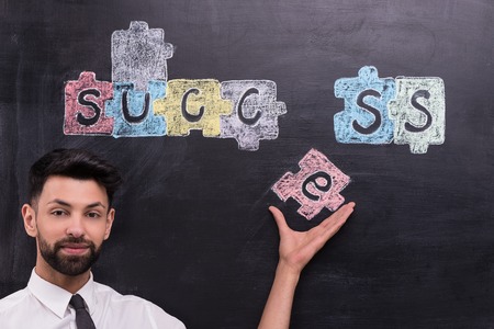 Young businessman on chalkboard background looking at camera and holding painted puzzle on chalkboard. Concept for successの写真素材