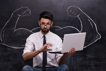 Funny picture of young businessman with laptop on chalkboard background. Two strong muscular arms painted on chalkboardの写真素材