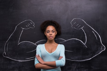 Funny picture of young afro-american woman on chalkboard background seriously looking at camera. Two strong muscular arms painted on chalkboardの写真素材