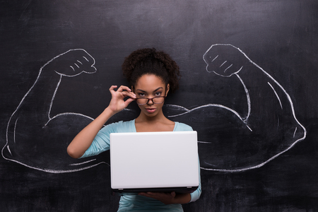 Funny picture of young afro-american woman with laptop on chalkboard background. Two strong muscular arms painted on chalkboardの写真素材