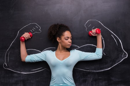 Funny picture of young afro-american woman with dumbbells on chalkboard background. Two strong muscular arms painted on chalkboardの写真素材