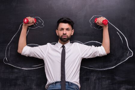 Funny picture of serious businessman with dumbbells on chalkboard background. Two strong muscular arms painted on chalkboardの写真素材