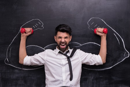 Funny picture of young businessman with dumbbells on chalkboard background. Two strong muscular arms painted on chalkboardの写真素材