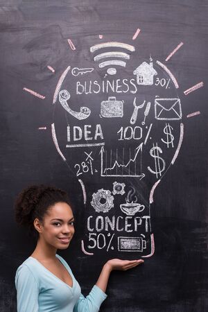 Smiling afro-american woman on chalkboard background looking at camera. Painted lightbulb with business ideas on chalkboard. Concept for successの写真素材