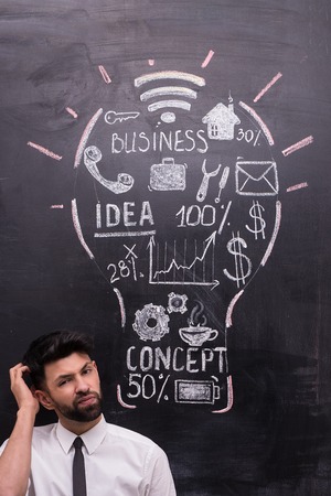 Thoughtful businessman on chalkboard background looking at camera. Painted lightbulb with business ideas on chalkboard. Concept for successの写真素材
