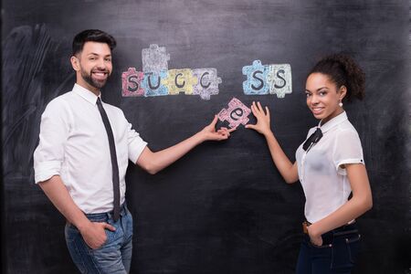 Young smiling afro-american woman and businessman on chalkboard background looking at camera and inserting painted puzzle are on chalkboard. Concept for successの写真素材