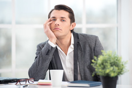 Young businessman is bored while working. Office interior with big window. Man looking at cameraの写真素材