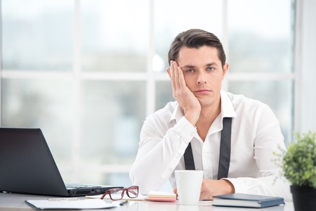 Young businessman is bored while working with computer. Office interior with big window. Man looking at cameraの写真素材