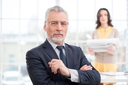Businessman looking at camera. His assistant holding a stack of papers. Office interior with big windowの写真素材