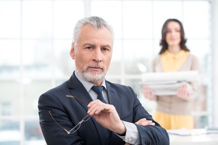 Businessman looking at camera. His assistant holding a stack of papers. Office interior with big windowの写真素材
