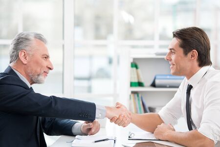 Two businessmen with documents and tablet computer. They shaking hands. Office interior with big windowの写真素材