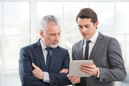 Two businessmen working on tablet computer and discussing something. Office interior with big windowの写真素材