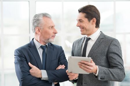 Two businessmen working on tablet computer and smiling. Office interior with big windowの写真素材