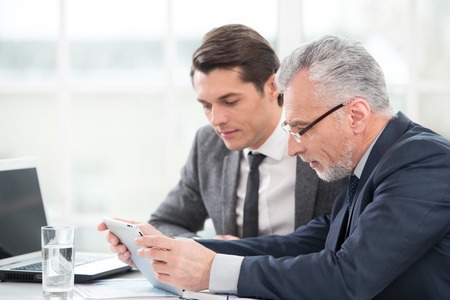 Two businessmen working with documents and tablet computer. Office interior with big windowの写真素材