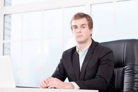 Young businessman seriously looking at camera and sitting at his black leather office chair. Office interior with windowの写真素材