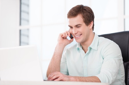 Young smiling businessman working with laptop and talking by phone while sitting at his black leather office chair. Office interior with windowの写真素材