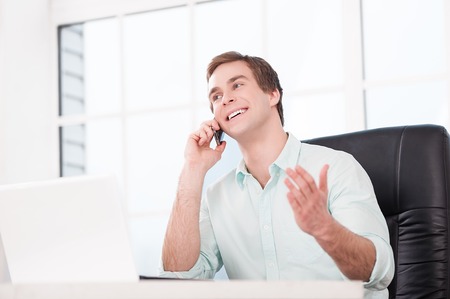 Young smiling businessman working with laptop and talking by phone while sitting at his black leather office chair. Office interior with windowの写真素材
