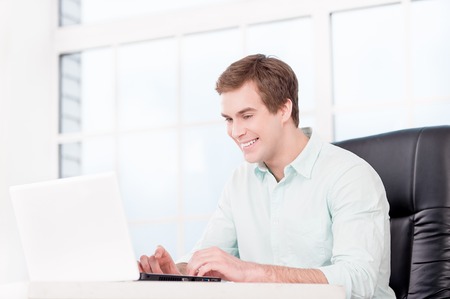 Young smiling businessman working with laptop and sitting at his black leather office chair. Office interior with windowの写真素材