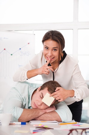 Young mixed race businesswoman gluing sticker on sleeping man while working day. Office interior with windowの写真素材