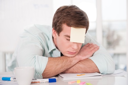 Young tired businessman with sticker on his forehead sleeping while working day. Office interior with windowの写真素材