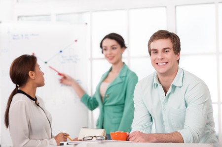 Young smiling businessman looking at camera. His creative team working with diagram on background. Office interior with windowの写真素材