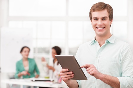 Young smiling businessman looking at camera and using tablet computer. His creative team working on background. Office interior with windowの写真素材