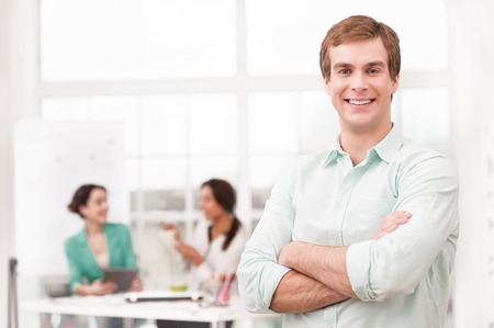 Young smiling businessman looking at camera. His creative team working on background. Office interior with windowの写真素材