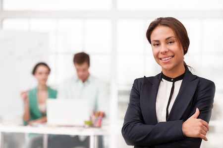 Young smiling mixed race businesswoman looking at camera. Her creative team working on background. Office interior with windowの写真素材