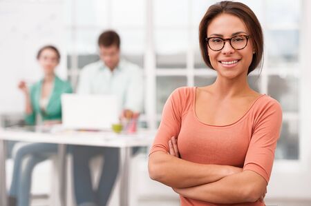 Young smiling mixed race businesswoman with glasses looking at camera. Her creative team working on background. Office interior with windowの写真素材