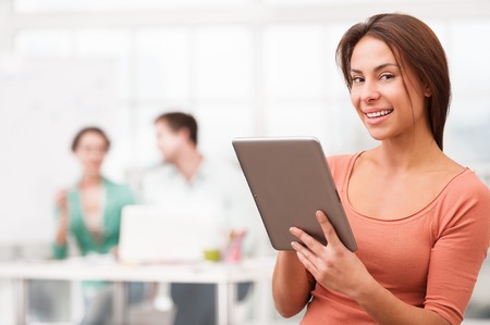 Young smiling mixed race businesswoman with tablet computer looking at camera. Her creative team working on background. Office interior with windowの写真素材