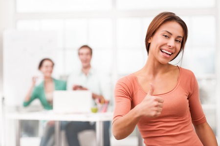 Young smiling mixed race businesswoman showing thumb up at camera. Her creative team working on background. Office interior with windowの写真素材