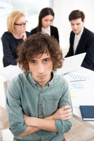 Photo of young casual businessman looking at camera with his business team on background. Office interior with window. Concept for teamworkの写真素材