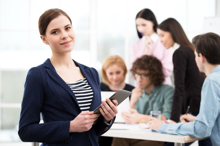 Businesswoman looking at camera and holding tablet computer with her business team on background. Office interior with window. Concept for teamworkの写真素材