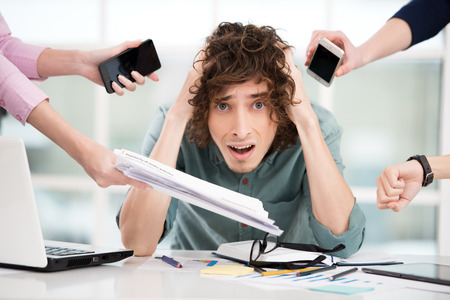 Sad young businessman looking at camera. Hands with gadgets around him. Office interior with window. Concept for overworkingの写真素材