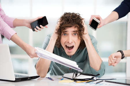 Shocked young businessman looking at camera. Hands with gadgets around him. Office interior with window. Concept for overworkingの写真素材