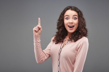 Excited young woman looking at camera and pointing up, standing on grey backgroundの写真素材