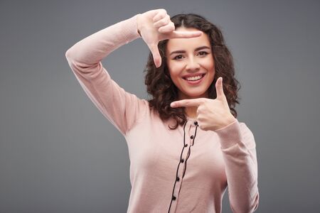 Beautiful young woman smiling and making hand frame, standing on grey backgroundの写真素材