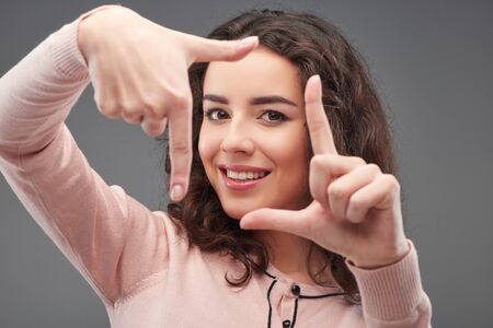 Close up portrait of young woman making hand frame, standing on grey backgroundの写真素材