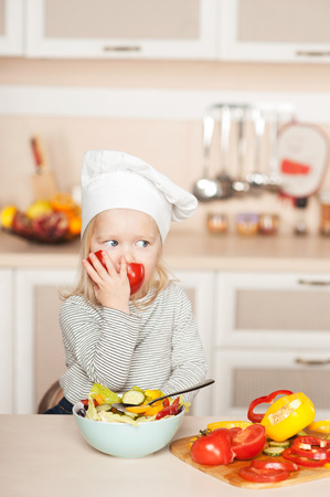 Little cute girl eating tomato while cooking salad. Kitchen interior. Concept for young kitchen handsの写真素材